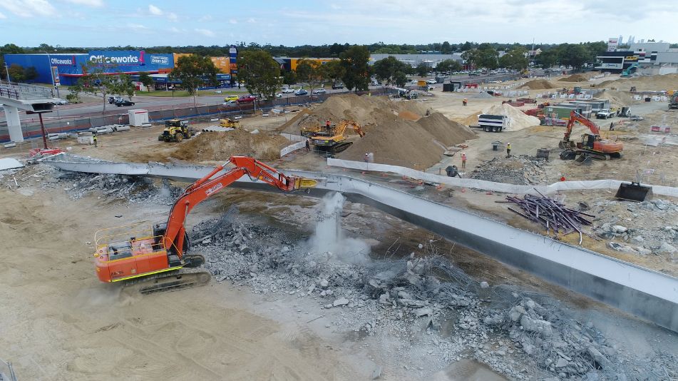 Wide shot of an orange excavator in Perth knocking down the carousel shopping center in Cannington, WA surrounded by sand and rubble.