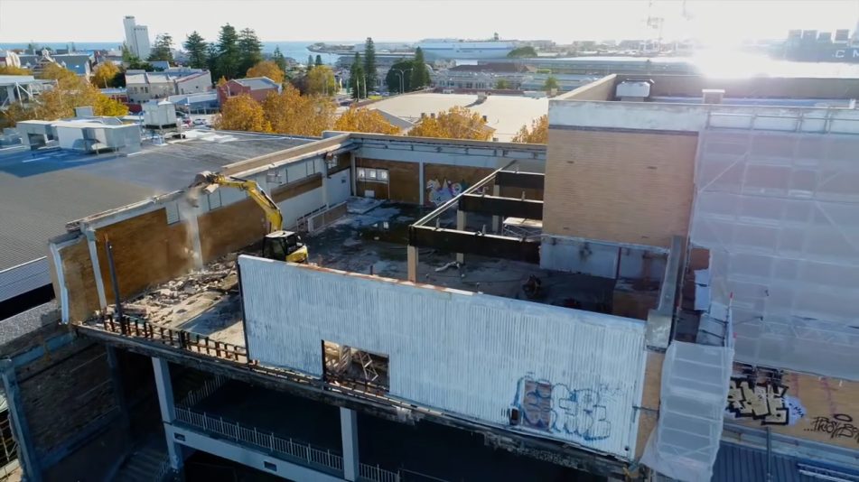 A yellow excavator inside a top story at the fremantle high rise building demolishing a part of the wall